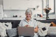 © Daniel - Elderly woman sits on a sofa at home smiling during a video call with family and friends. She enjoys connecting and sharing happy moments through modern technology.