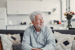 © Daniel - Elderly man sits on a sofa at home looking outside the window. He feels calm and reflective while enjoying a serene moment in his daily life.