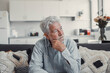© Daniel - Senior man sits alone on a sofa looking outside with a sad expression. He seems stressed and reflective while dealing with personal concerns at home.