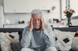 © Daniel - Mature man sits on a sofa holding his head in discomfort. He experiences headache and fatigue while coping with stress and illness at home.