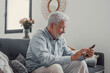 © Daniel - Senior man sits on a sofa smiling while using his smartphone. He enjoys browsing, buying, and connecting with the digital world at home.