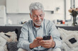 © Daniel - Senior man sits on a sofa smiling while using his smartphone. He enjoys browsing, buying, and connecting with the digital world at home.