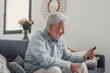 © Daniel - Senior man sits on a sofa smiling while using his smartphone. He enjoys browsing, buying, and connecting with the digital world at home.
