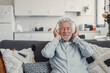 © Daniel - Elderly man sits on a sofa at home enjoying music with headphones. He feels relaxed, happy, and carefree while immersing himself in sound and leisure.