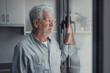 © Daniel - Elderly man looks out the window with concern. He appears thoughtful and worried about his life and surroundings while standing at home.