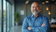© miss irine - Bald man with gray beard smiles, crosses arms in office setting. Mature professional looks confident, experienced, ready for business meeting. Positive executive poses near window, blurred background.