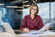 © Liubomir - Businesswoman with short blonde hair and glasses carefully reviewing paperwork and writing on a clipboard while working on her laptop at an office desk