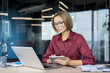 © Liubomir - Confident businesswoman with eyeglasses working diligently on a laptop and holding a digital tablet at a light wooden desk, focusing on tasks in a contemporary office environment