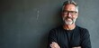 © Vadym - Mature man with grey beard and glasses smiles. He wears a black t-shirt and has crossed arms. Person stands indoors with a dark textured background. Man looks confident and relaxed.