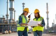© Pete - Two women in hard hats and safety vests confer over blueprints at industrial plant. One woman speaks into walkie-talkie, coordinating with team members on site. Refinery structures loom in background.