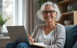 © zhao - Happy stylish mature old woman remote working from home distance office on laptop taking notes. Smiling 60s middle aged business lady using computer watching webinar sit on couch writing in notebook.