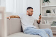 © SHOTPRIME STUDIO - Relaxed man sitting on a sofa, engaging with his smartphone, in a modern and bright living room with minimalistic decor and greenery. Perfect for lifestyle themes in 2025.