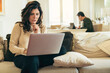 © Lomb - Middle aged woman working from home using laptop in living room with partner in background