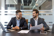 © Liubomir - Two smiling adult businessmen, one holding documents and gesturing while talking, collaborating and exchanging ideas at a modern office conference table with laptops and a tablet