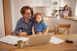 © Stockphotodirectors - A father is seated at a table with his young child, both focused on a laptop. In the background, a mother prepares food in a bright kitchen, creating a warm family atmosphere.