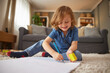 © Stockphotodirectors - A child sits on a soft rug in a cozy living room, happily drawing with a pencil and holding a yellow eraser, enjoying quality family time indoors on a sunny day.