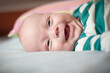 © guruXOX - A baby wearing a striped green and white shirt is lying on their back on a light blue bedspread. The baby is smiling with their mouth open and looking at the camera.