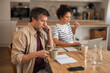 © Jelena - Thoughtful Couple At Kitchen Table Working From Home With Laptop And Notebooks
