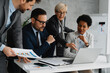 © Novak - Multi-ethnic team of colleagues sitting together at a conference table with a laptop and documents, discussing project strategy