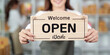© Natee Meepian - Open for business. Woman holding 'Welcome Open' sign in retail environment.
