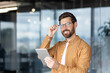 © Liubomir - Confident young businessman in a modern office holding a tablet, adjusting his glasses and smiling at the camera friendly, professional portrait conveying tech, innovation and success