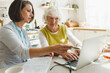 © shurkin_son - Senior lady learning to use laptop and online applications under assistance of female social support volunteer teaching retired person cyber technology at home, explaining and instructing