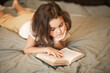 © zulfiska - A thoughtful young girl lies on her stomach on a cozy bed, deeply engrossed in reading an open book, resting her chin on her hand.