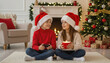 © Svetlana - Two children wearing Santa hats sitting together by Christmas tree