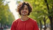 © Maryna - Young man with curly brown hair wears a red shirt outdoors. He smiles warmly standing in a park with blurred trees and buildings in background. Casual portrait shot.
