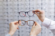 © st.kolesnikov - Hands comparing two eyeglass frames in optical store choosing eyewear for vision correction