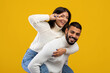 © Prostock-studio - A young couple enjoys a lighthearted moment with smiles, the woman playfully posing on the man's back in cozy sweaters. The vibrant yellow backdrop adds joy to the scene.