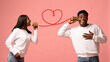 © Prostock-studio - Two joyful African American lovers communicate through a tin can phone, sharing words of love and fun on Valentine's Day, with a bright pink studio background enhancing the festive mood.