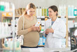© JackF - Interested pregnant woman reading label on stretch mark cream bottle, focusing on natural ingredients, while friendly female pharmacist providing guidance on safe skincare in background at drugstore