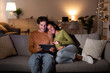 © Prostock-studio - Happy couple sits close on their couch, smiling as they watch a movie on their laptop. It is nighttime, and a warm ambiance fills the room with soft lighting.