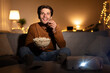 © Prostock-studio - A young man smiles as he watches a movie using a projector in his cozy living room. He enjoys a bowl of popcorn while relaxing on the sofa during a weekend evening.