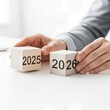 © Alima.Std - Hands Arranging Wooden Blocks with Years 2025 and 2026, Business Planning and Future Transition Concept.