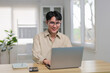 © amnaj - Young Asian man wearing glasses smiling and working on a laptop at a desk in a contemporary, bright office setting
