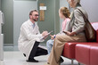 © pressmaster - Caucasian male doctor kneeling and speaking with Caucasian girl child while senior Caucasian woman sitting nearby in waiting area, doctor holding clipboard and pen, medical consultation scene