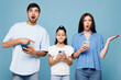 © ViDi Studio - Young shocked parents mom dad with child kid daughter girl wearing casual clothes hold in hand use peek on mobile cell phone isolated on plain blue background. Family day parenthood childhood concept.