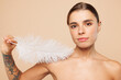 © ViDi Studio - Beautiful half naked topless young woman 20s with nude make up touching shoulder with feather isolated on plain beige color background studio portrait Skin care healthcare cosmetic procedures concept