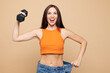 © ViDi Studio - Young woman wear orange casual clothes hold dumbbell show loose denim pants after weight loss isolated on plain pastel beige background. Proper nutrition healthy fast food unhealthy choice concept