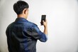 © yashis007 - Man in Denim Jacket Holding and Looking at Smartphone Against White Background, Back View