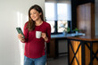 © NDABCREATIVITY - Pregnant woman enjoying and smelling cup of coffee while scrolling through her phone in the morning