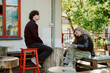 © m-art - Two friends sit comfortably in a sunlit outdoor cafe, sharing a moment. One writes while the other gazes thoughtfully, surrounded by greenery and charming decor.