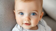 © A2Z AI  - Close-up portrait of a baby with big blue eyes and fair skin. The baby is wearing a light-colored shirt, and is looking directly at the viewer, with a slight smile. The background is a neutral color.