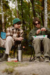 © m-art - Two young men sit on camping chairs by a serene forest stream. One is preparing bait while the other focuses on fishing. Lush trees surround them, creating a calm atmosphere for their adventure.