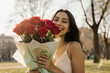 © sumon78 - Happy young woman smiling holds beautiful red rose bouquet gift. She enjoys romantic present in an outdoor park during sunny day, feeling joyful and appreciated