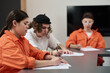 © pressmaster - Group of young adults sitting at table completing written assignments in classroom setting, wearing orange prison uniforms, participating in prison education program
