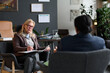 © TrueFrame Collective - Senior Caucasian woman holding clipboard listening attentively to adult woman during therapy session in office setting, both seated facing each other, discussing mental health concerns