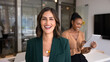 © fizkes - Happy beautiful Hispanic businesswoman posing for head shot in office workspace with diverse colleagues working behind, looking at camera, smiling. Banner portrait of confident female business leader